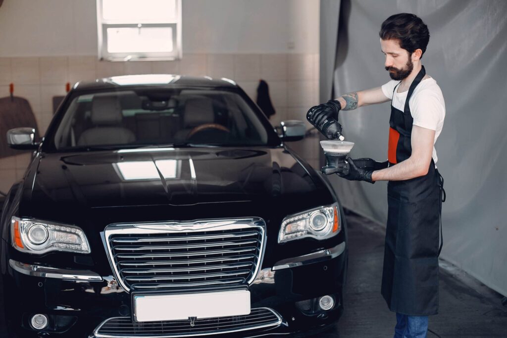 Male car detailer in a white shirt and black apron pouring a ceramic coating or polishing product from a bottle into an applicator cup while standing in front of a shiny black luxury sedan.
