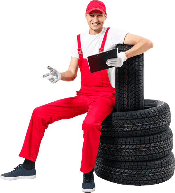 Friendly, smiling male mechanic in red overalls, white shirt, and a red cap, sitting on a stack of four new winter or all-season tires while holding a clipboard; isolated on a grey background.
