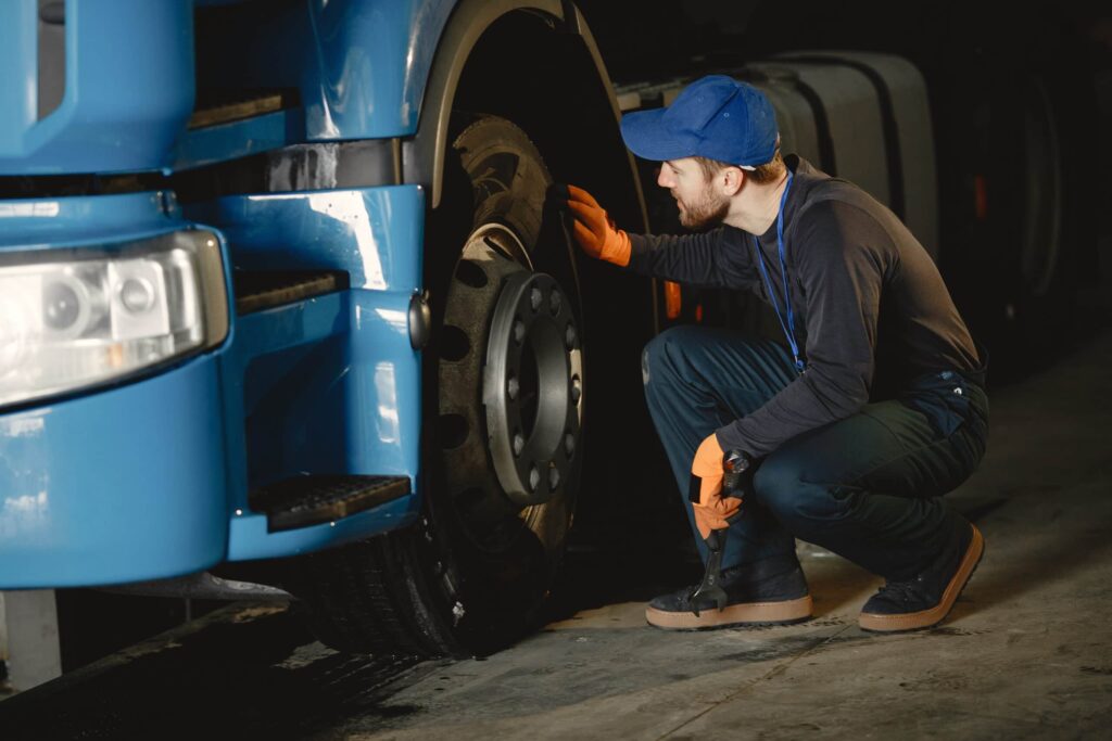 A professional mechanic in a blue cap and orange gloves inspecting about a large blue semi-truck tire in a service garage.