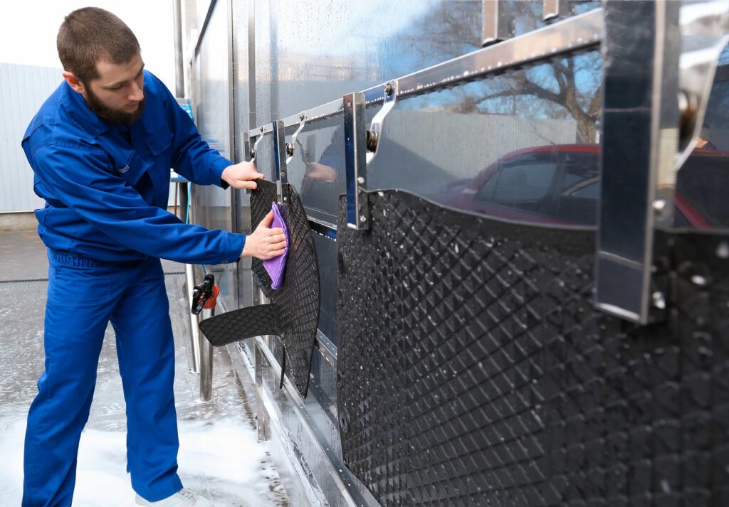 A professional car wash worker in blue coveralls uses a purple microfiber cloth to wipe down and dry black rubber car mats that are hung on a stainless steel rack.