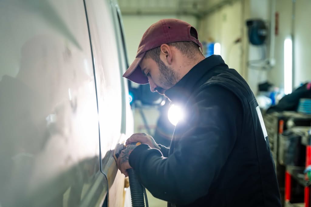 A professional detailer carefully operating a dual action sander or polishing tool along the side panel of a white vehicle under bright studio lighting inside a Los Angeles, California auto repair shop.