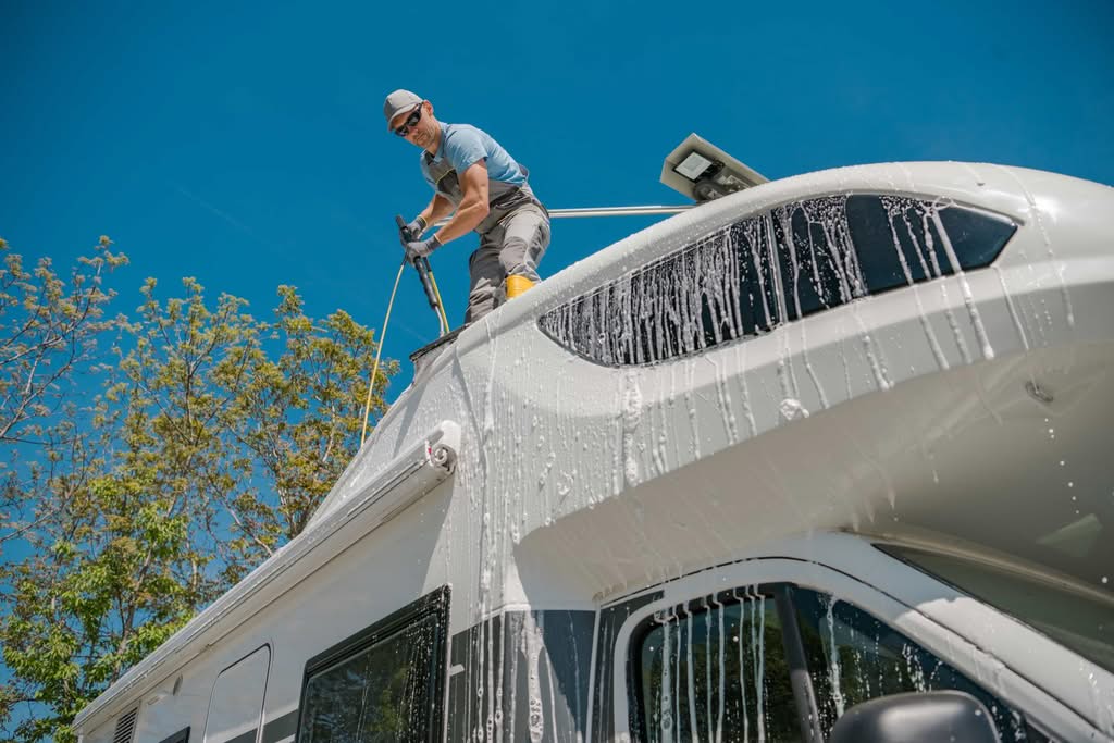 A professional detailer standing safely on top of a large white motorhome roof using a wash wand and soap to deep clean the fiberglass surface during an RV gelcoat restoration service in Ocala, Florida.